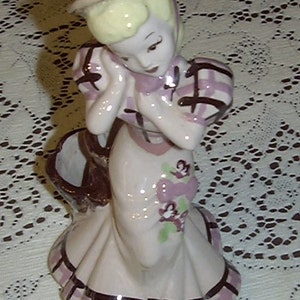 May include: A porcelain figurine of a girl wearing a pink and white dress with a brown basket. She has blonde hair and is wearing a brown hat. The figurine is decorated with pink flowers.