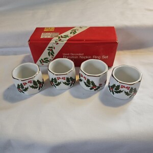 May include: A set of four white porcelain napkin rings with gold trim, decorated with green holly leaves and red berries. The napkin rings are displayed in front of a red box with the text "Hand Decorated Porcelain Napkin Ring Set".