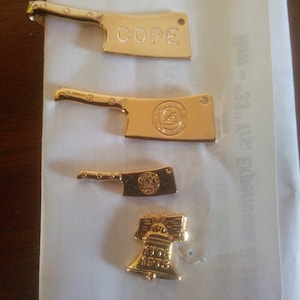 May include: Four gold-colored metal charms, including a butcher knife with the word "COPE" engraved on it, a butcher knife with a logo, a butcher knife with a logo, and a Liberty Bell charm with the year "1976" engraved on it.
