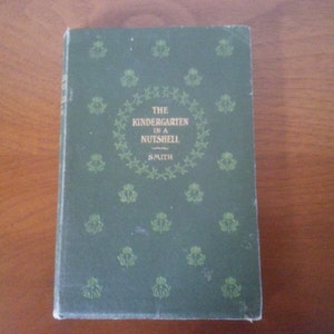 May include: A green book with a gold title that reads "The Kindergarten in a Nutshell Smith". The cover is decorated with a repeating pattern of small floral designs.
