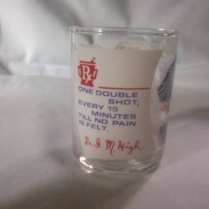 May include: A clear glass shot glass with a white label that reads "R ONE DOUBLE SHOT, EVERY 15 MINUTES TILL NO PAIN IS FELT. Ph.d. M. High."
