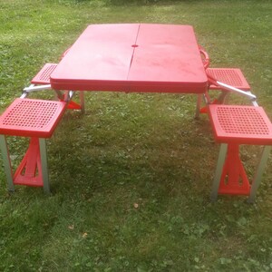 May include: Red folding picnic table with four red folding chairs. The table and chairs are made of plastic and metal.