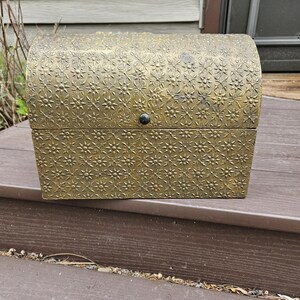 May include: A decorative, antique-style chest with a domed lid. The chest is a golden color and features an embossed floral pattern. It has a small, round, black clasp on the front.