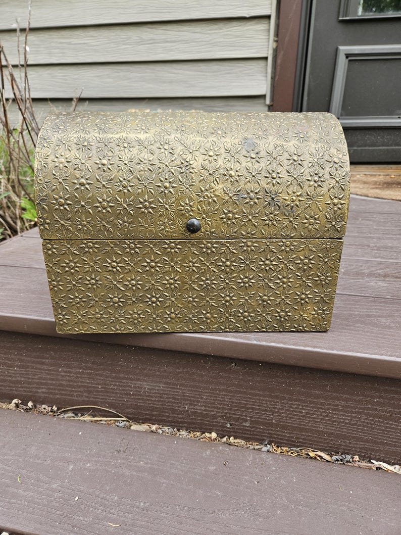 May include: A decorative, antique-style chest with a domed lid. The chest is a golden color and features an embossed floral pattern. It has a small, round, black clasp on the front.