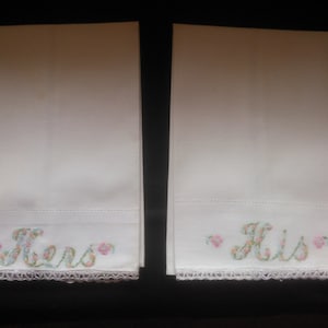 May include: Two white hand towels with embroidered floral lettering. The towel on the left says "Hers" and the towel on the right says "His".