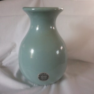 May include: A light blue ceramic vase with a round body and a wide mouth. The vase has a small, round sticker on the bottom with the word "Mia" printed on it.