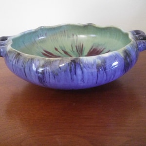 May include: A ceramic bowl with two handles, featuring a green, purple, and black glaze. The bowl has a unique, organic shape and a glossy finish.