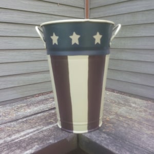 May include: A metal planter with a striped design in brown and cream with a blue band and white stars at the top.