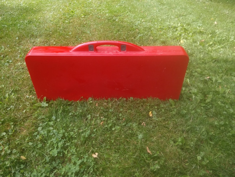 May include: A red folding table and chairs set in a closed position. The set has a carrying handle and is designed for outdoor use.