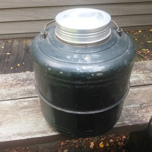May include: A vintage green metal insulated beverage container with a silver lid. The container has a rounded shape and a slightly textured surface.