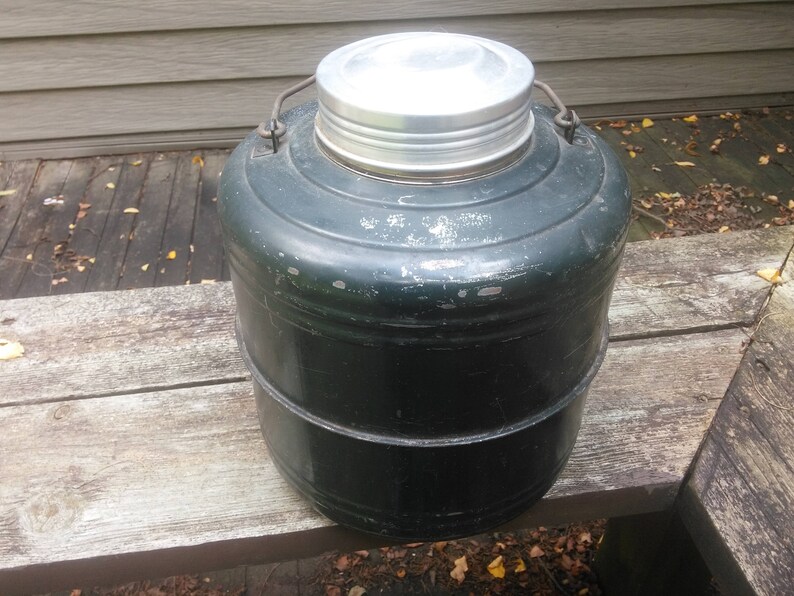 May include: A vintage green metal insulated beverage container with a silver lid. The container has a rounded shape and a slightly textured surface.