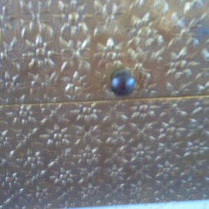 May include: Close-up of a gold-colored box with a floral pattern. The box has a dark, round knob. The floral pattern is embossed across the surface of the box. The box appears to be made of a metallic material.