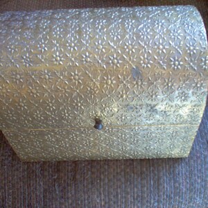 May include: A vintage, gold-toned chest with a floral pattern embossed across its curved lid and sides. The small chest has a dark, round knob for opening and closing. The chest is likely used for storing jewelry or small keepsakes.