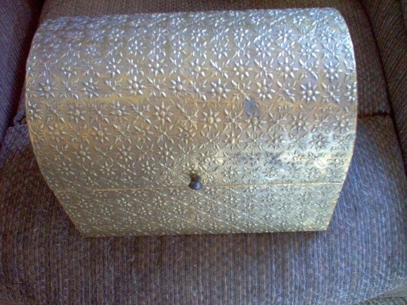 May include: A vintage, gold-toned chest with a floral pattern embossed across its curved lid and sides. The small chest has a dark, round knob for opening and closing. The chest is likely used for storing jewelry or small keepsakes.