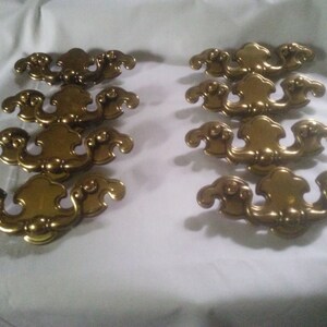 May include: Set of ten gold-toned cabinet drawer pulls with a decorative, ornate design. Each pull has a curved shape with a raised center and intricate details.