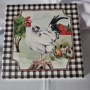 May include: A white rooster with a red comb stands on a tree stump in front of a red barn. The rooster is surrounded by green foliage and three smaller chickens. The image is framed with a black and white checkered border.