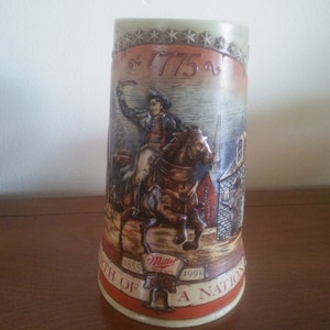 May include: A ceramic beer stein with a brown, white, and red design. It features a depiction of a man on horseback with the text "1775" and "Birth of a Nation" on the stein.