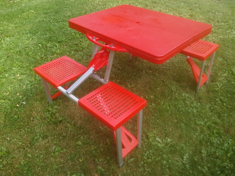 May include: Red folding picnic table with three red folding chairs. The table and chairs are made of plastic and metal.