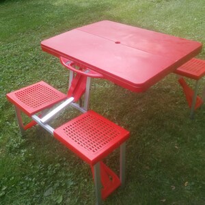 May include: A red folding picnic table with four red folding chairs. The table and chairs are made of plastic and metal.