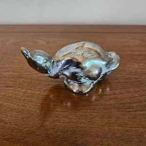 May include: A silver-colored ceramic turtle figurine with a distressed white paint finish. The turtle is in a running pose with its head raised.