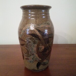 May include: A handcrafted ceramic vase with a speckled brown and tan glaze. The vase features abstract dark brown and green painted designs. The top has a slightly flared rim with horizontal grooves.