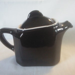May include: A black ceramic teapot with a white rim and a lid. The teapot has a square body and a curved spout.