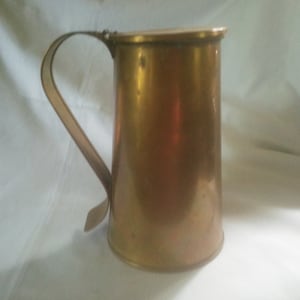 May include: A gold-colored metal pitcher with a handle. The pitcher has a rounded body and a slightly flared lip.