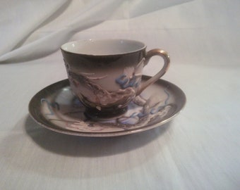 May include: A white teacup and saucer with a black and white dragon design. The cup has a gold rim and the saucer has a gold trim.