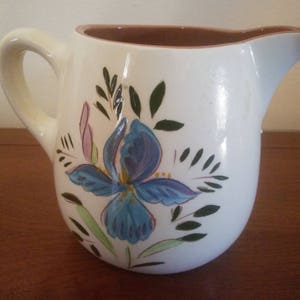 May include: White ceramic pitcher with a blue and purple floral design. The pitcher has a brown rim and a handle.