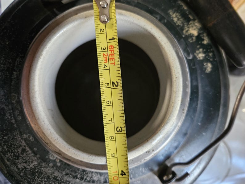 May include: Close-up of a black metal container with a white ceramic lid. A yellow measuring tape is placed next to the container, showing a measurement of 3 inches.
