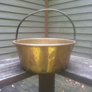 May include: A large, round, brass pot with a black metal handle. The pot is sitting on a wooden surface.