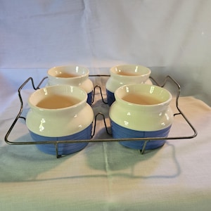 May include: Four ceramic pots with a white top, blue base, and a light brown interior, arranged on a metal rack. The pots have a rounded shape and are evenly spaced. The background is a white cloth.