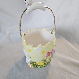 May include: A ceramic Easter basket shaped like a cracked egg, painted yellow with yellow polka dots. A white bunny is perched on the handle. The basket is decorated with flowers and another bunny.