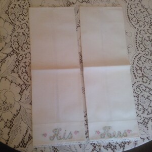 May include: Two white hand towels with embroidered lace trim and the words "His" and "Hers" in pink and green lettering.