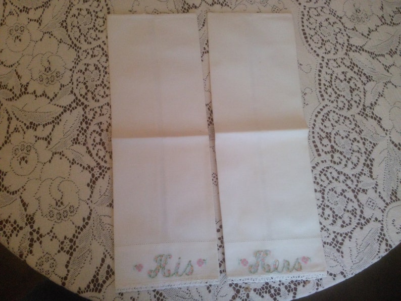 May include: Two white hand towels with embroidered lace trim and the words "His" and "Hers" in pink and green lettering.