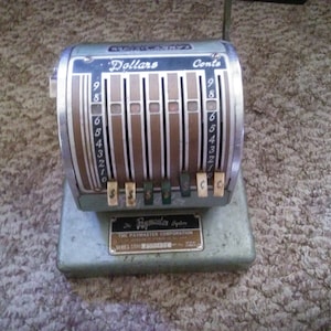 May include: A vintage green metal cash register with a silver faceplate. The register has a series of numbered dials for dollars and cents. The register is labeled "The Paymaster System" and "Series 2500".