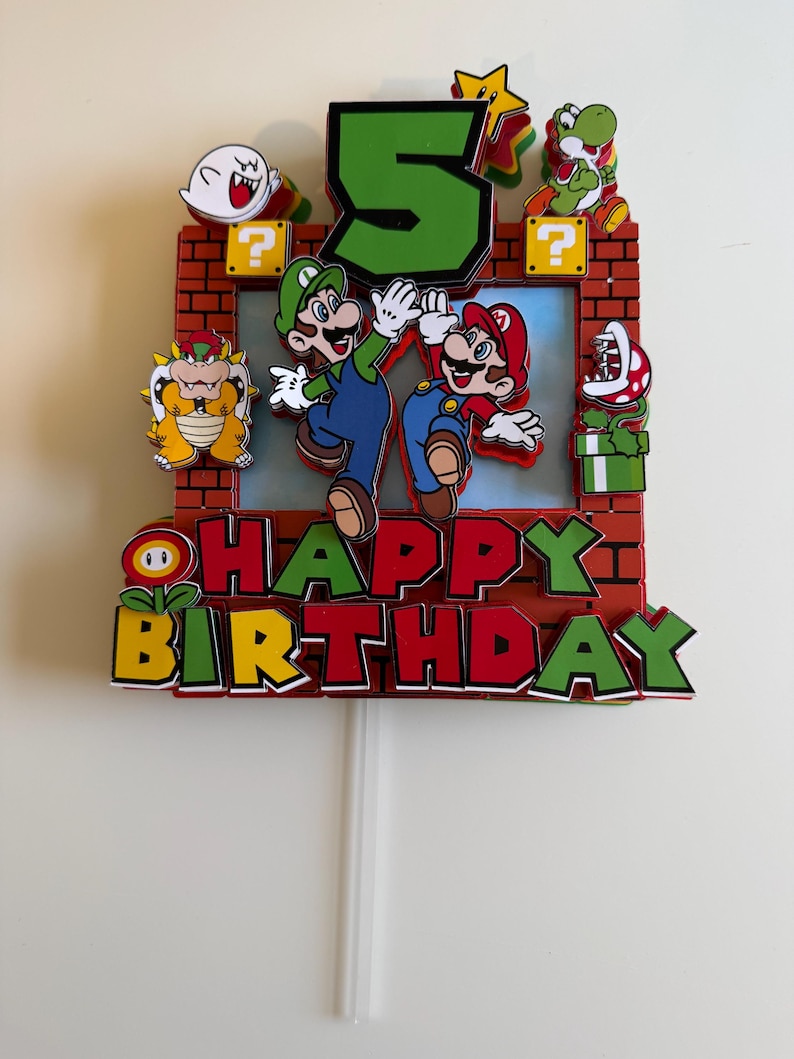 Op de afbeelding: Een meerlaagse papieren taarttopper met een groot groen cijfer 5, de personages Mario en Luigi, Bowser, Yoshi, een Boo-geest en een Piranha-plant. Het ontwerp bevat een rood bakstenen frame met gele vraagtekenblokken en de tekst HAPPY BIRTHDAY.