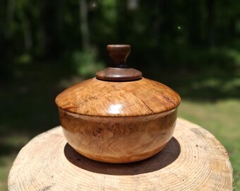 May include: A round wooden bowl with a polished, light brown finish featuring natural wood grain patterns and a smooth texture. The bowl includes a matching lid with a dark brown, knob-shaped handle centered on top, creating a cohesive look.