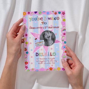A colorful birthday invitation is held between two hands. The card has pastel stripes and a border filled with playful stickers like stars, hearts, smiley faces, rainbows, strawberries, and flowers. In the center, a round black‑and‑white photo of a smiling child sits beside a cute birthday cake illustration. The bright, crafty design creates a fun, whimsical celebration feel.