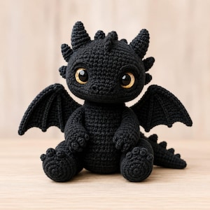 Può includere: Un peluche a forma di drago lavorato all'uncinetto in nero, con un corpo in filato testurizzato, due piccole ali, una coda spinata e grandi occhi rotondi color ambra. La creatura è raffigurata in posizione seduta con cuciture visibili.