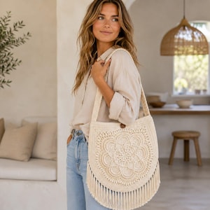 May include: A cream-colored crochet shoulder bag featuring a large, intricate circular mandala pattern on the front, a single wide strap, and long fringe detailing along the bottom edge, secured with a small brown button at the top center.