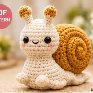 May include: A crochet snail toy featuring a cream-colored body with two antennae topped with mustard yellow spheres, a large mustard yellow spiral shell, black bead eyes, and pink stitched cheeks. The image includes the text PDF PATTERN.