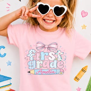 May include: A light pink t-shirt featuring a graphic print with a large pink bow above the text "first grade" in blue and pink patterned letters. Below the text is a blue pencil graphic with the name "Amelia" written in pink script.