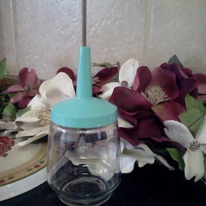 May include: A vintage glass food mill with a teal colored top and a metal rod. The mill is sitting on a black surface with a floral arrangement in the background.