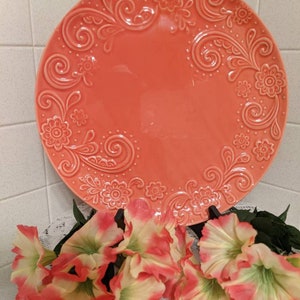 May include: A large, round, coral-colored ceramic serving platter with an embossed floral design. The platter has a raised rim and a smooth, glossy finish.