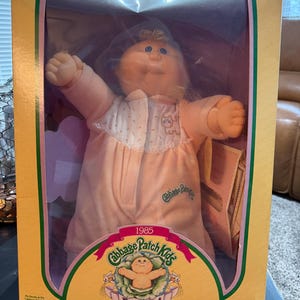 May include: A vintage Cabbage Patch Kids Preemie doll in its original box. The doll is wearing a pink outfit with a white bib. The box is yellow with green and pink accents and features the text "Cabbage Patch Kids" and "Preemie!"