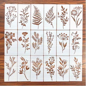 May include: A collection of botanical stencils featuring various floral and foliage designs. Each stencil showcases a different plant, including ferns, flowers, and sprigs, in a brown color against a white background. The stencils are arranged on a wooden surface.