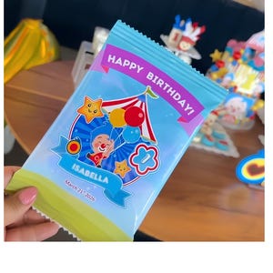 May include: A light blue rectangular party favor bag featuring a circus tent graphic, a cartoon character holding balloons, and the text "HAPPY BIRTHDAY!", "ISABELLA", and "March 21st 2026" printed on the front with a green bottom border.