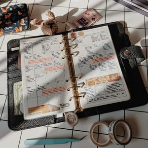 May include: A black leather six-ring binder planner open to a monthly grid calendar with handwritten notes, pink highlighter strips, and the text "Kuns" and "Ja!". Accessories include a small crochet animal, two rolls of washi tape, and tweezers.
