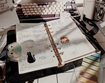 May include: An open six-ring planner with grid paper, featuring a black cat sticker, a cartoon character sticker, and a pastel frog-shaped page marker. The planner rests on a black textured cover, surrounded by a mechanical keyboard and sticky notes.